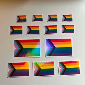 May include: A collection of Pride flag stickers in various sizes. The stickers feature the Progress Pride flag design with rainbow stripes, a chevron of colors, and a white border. Some stickers have a holographic finish.