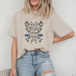 May include: A beige short-sleeve t-shirt featuring a central graphic with the Latin text Audentes fortuna iuvat, surrounded by illustrations of crossed swords, green vines, red flowers, and blue leaves, paired with light blue distressed denim jeans.