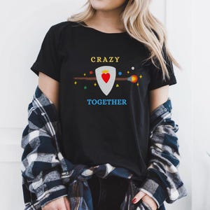 Könnte beinhalten: Ein schwarzes Kurzarm-T-Shirt mit dem gelben Schriftzug CRAZY über einer weißen Schildgrafik mit rotem Herz und Krone, einem braunen Zauberstab und dem blauen Schriftzug TOGETHER darunter, umgeben von kleinen bunten geometrischen Formen und Sternen.