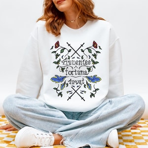May include: A white long-sleeve sweatshirt featuring a central graphic with the Latin text Audentes fortuna iuvat. The design includes two crossed black swords, red flowers, green leaves, and blue foliage elements arranged in a symmetrical pattern.