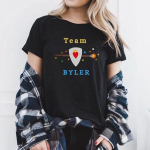 以下が含まれることがあります： 「Team BYLER」の文字が黄色と青でプリントされた黒いTシャツ。デザインには赤いハートの盾と、カラフルなアクセントが付いた様式化された矢が含まれています。Tシャツは半袖です。