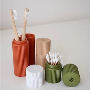 May include: A collection of cylindrical, vertically ribbed containers in terracotta, beige, white, and olive green. Two bamboo toothbrushes are inside the terracotta holder, while white cotton swabs are stored in the olive green container.