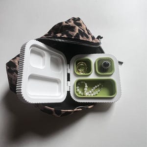 May include: A white and green jewellery organiser with a leopard print bag. The organiser is open, revealing compartments holding a gold ring, a pearl necklace, and a black gemstone ring. The bag is black with a leopard print.
