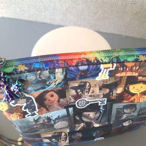 May include: A colorful zippered pouch with a rainbow zipper and a variety of images printed on it. The images include characters from the movie "Coraline" and a key with a film reel design.