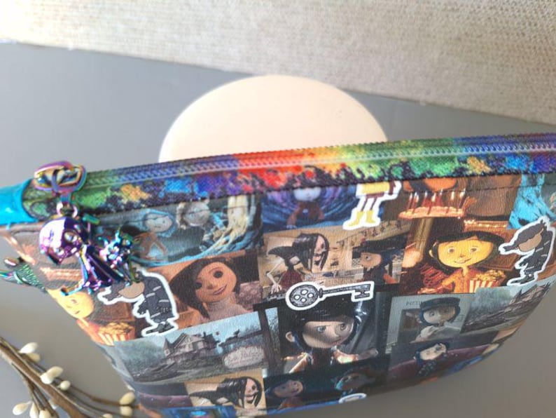 May include: A colorful zippered pouch with a rainbow zipper and a variety of images printed on it. The images include characters from the movie "Coraline" and a key with a film reel design.