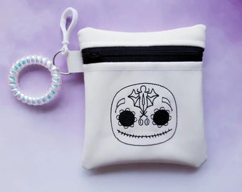 May include: White zippered pouch with a black zipper and a black embroidered design of a skull with a bat on the top. The pouch has a white iridescent spiral hair tie attached to it.