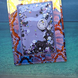 Peut inclure: Une carte shaker faite &agrave; la main avec un fond violet et une fen&ecirc;tre transparente remplie de confettis en forme de cr&acirc;ne et de paillettes. La carte pr&eacute;sente un motif peau de serpent orange, bleu et jaune. Des formes de cr&acirc;nes et de c&oelig;urs appliqu&eacute;es sont visibles.