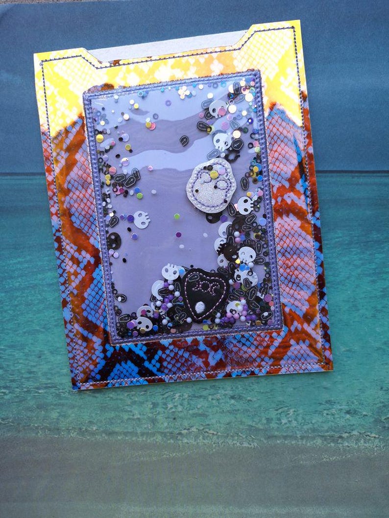 Peut inclure: Une carte shaker faite &agrave; la main avec un fond violet et une fen&ecirc;tre transparente remplie de confettis en forme de cr&acirc;ne et de paillettes. La carte pr&eacute;sente un motif peau de serpent orange, bleu et jaune. Des formes de cr&acirc;nes et de c&oelig;urs appliqu&eacute;es sont visibles.