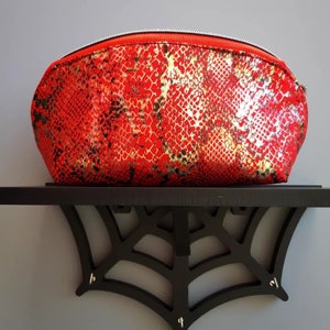 May include: A red cosmetic bag with a snakeskin pattern in gold and black. The bag has a curved shape and a silver zipper. The bag is sitting on a black shelf with a spiderweb design.