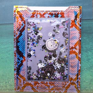May include: A decorative, rectangular card holder with a clear, shaker-style center filled with skull-shaped confetti. The holder has a snakeskin pattern in orange, blue, and purple. The center features a skull and a heart.