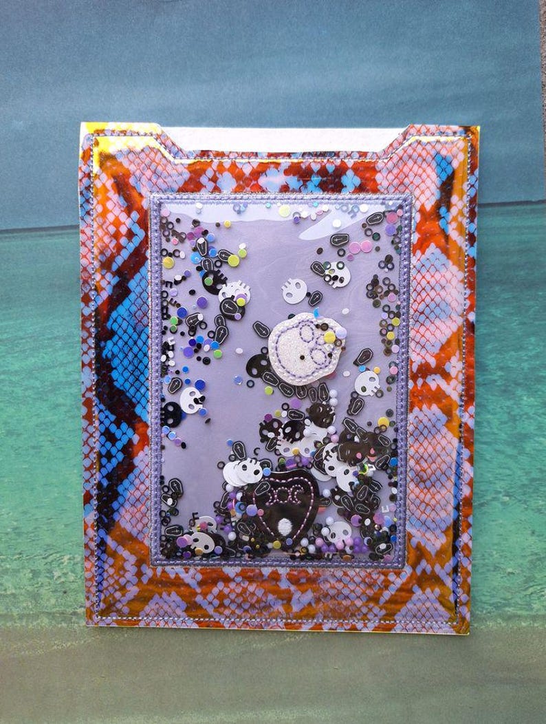 Peut inclure: Un porte-cartes rectangulaire d&eacute;coratif avec un centre transparent de style shaker rempli de confettis en forme de cr&acirc;ne. Le support pr&eacute;sente un motif peau de serpent orange, bleu et violet. Le centre pr&eacute;sente un cr&acirc;ne et un c&oelig;ur.