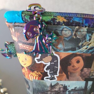 May include: A colorful zippered pouch with a variety of images from the movie "Coraline" printed on it. The pouch has a silver zipper and a silver charm attached to the zipper pull. The charm is a silhouette of the main character from the movie, Coraline.