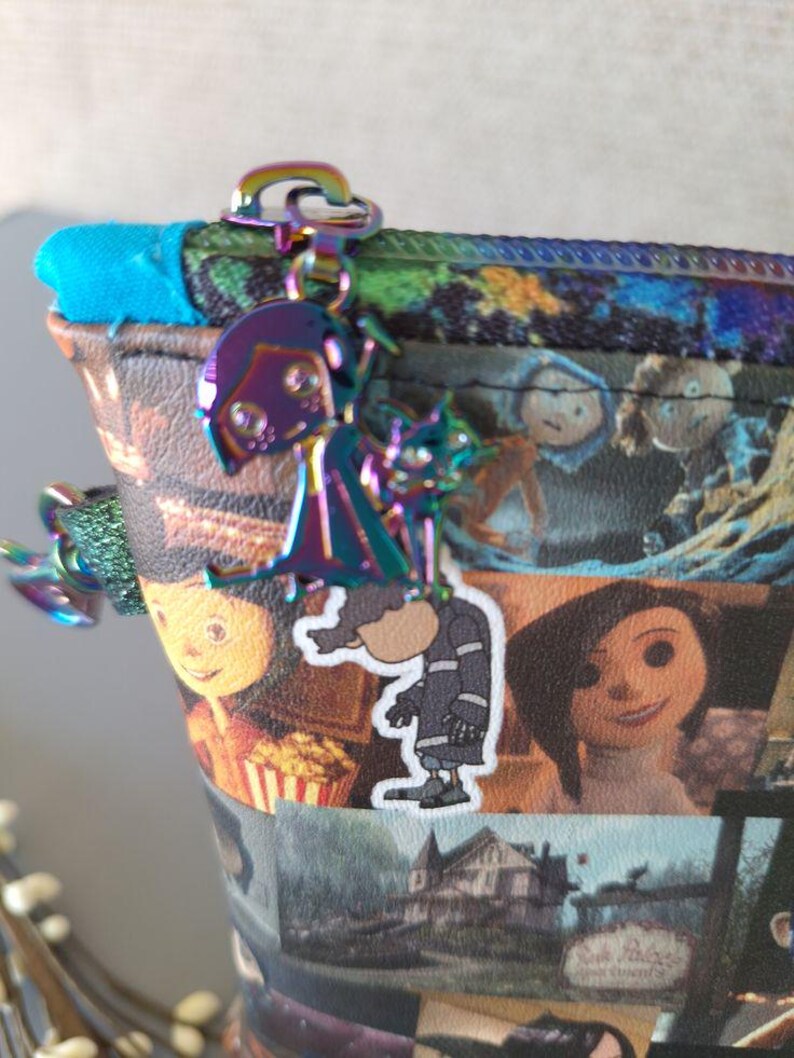 May include: A colorful zippered pouch with a variety of images from the movie "Coraline" printed on it. The pouch has a silver zipper and a silver charm attached to the zipper pull. The charm is a silhouette of the main character from the movie, Coraline.