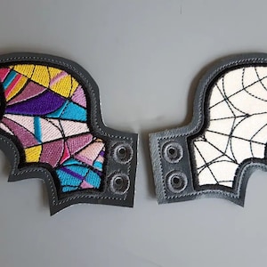May include: A pair of gray felt bat wings with embroidered designs. One wing has a colorful geometric pattern, while the other has a white spiderweb design.