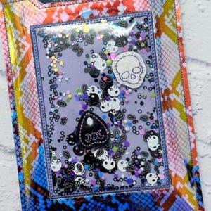 Peut inclure: Une housse de liseuse color&eacute;e avec un motif peau de serpent bleu, orange et jaune. Le panneau avant transparent pr&eacute;sente un design &agrave; secouer avec des embellissements sur le th&egrave;me des cr&acirc;nes et des planches Ouija. Le texte "SARAH J. MAAS" est visible.