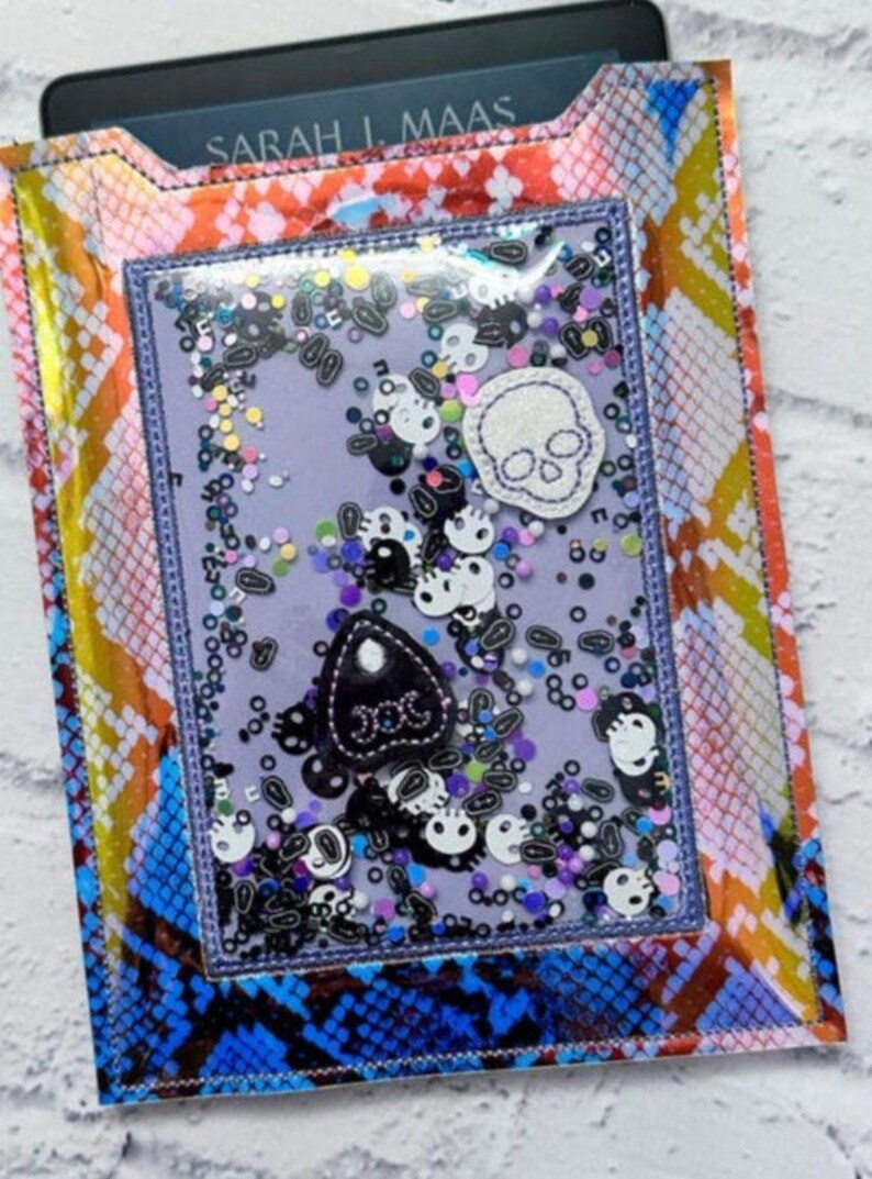 Peut inclure: Une housse de liseuse color&eacute;e avec un motif peau de serpent bleu, orange et jaune. Le panneau avant transparent pr&eacute;sente un design &agrave; secouer avec des embellissements sur le th&egrave;me des cr&acirc;nes et des planches Ouija. Le texte "SARAH J. MAAS" est visible.