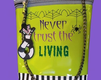 Può includere: Una borsa a tracolla a righe verdi e nere con una tracolla a catena. La borsa ha una chiusura a zip e presenta un design ricamato che dice "Never trust the living" con un ragno e una ragnatela sopra il testo. Un personaggio bianco e nero con una lingua verde è attaccato alla tracolla a catena.