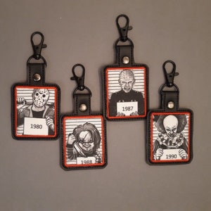 May include: Four black leather keychains with red stitching and a white background. Each keychain features a different horror movie character with a mugshot style image and the year the movie was released. The characters are Jason Voorhees from Friday the 13th (1980), Freddy Krueger from A Nightmare on Elm Street (1988), Pinhead from Hellraiser (1987), and Pennywise from IT (1990).