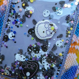 Peut inclure: Un shaker rectangulaire transparent avec un fond violet rempli de confettis en forme de cr&acirc;nes et de cercueils. Un &eacute;cusson de cr&acirc;ne blanc avec des coutures violettes est au centre. Le shaker est bord&eacute; d'une bordure violette et repose sur une surface color&eacute;e &agrave; motifs.