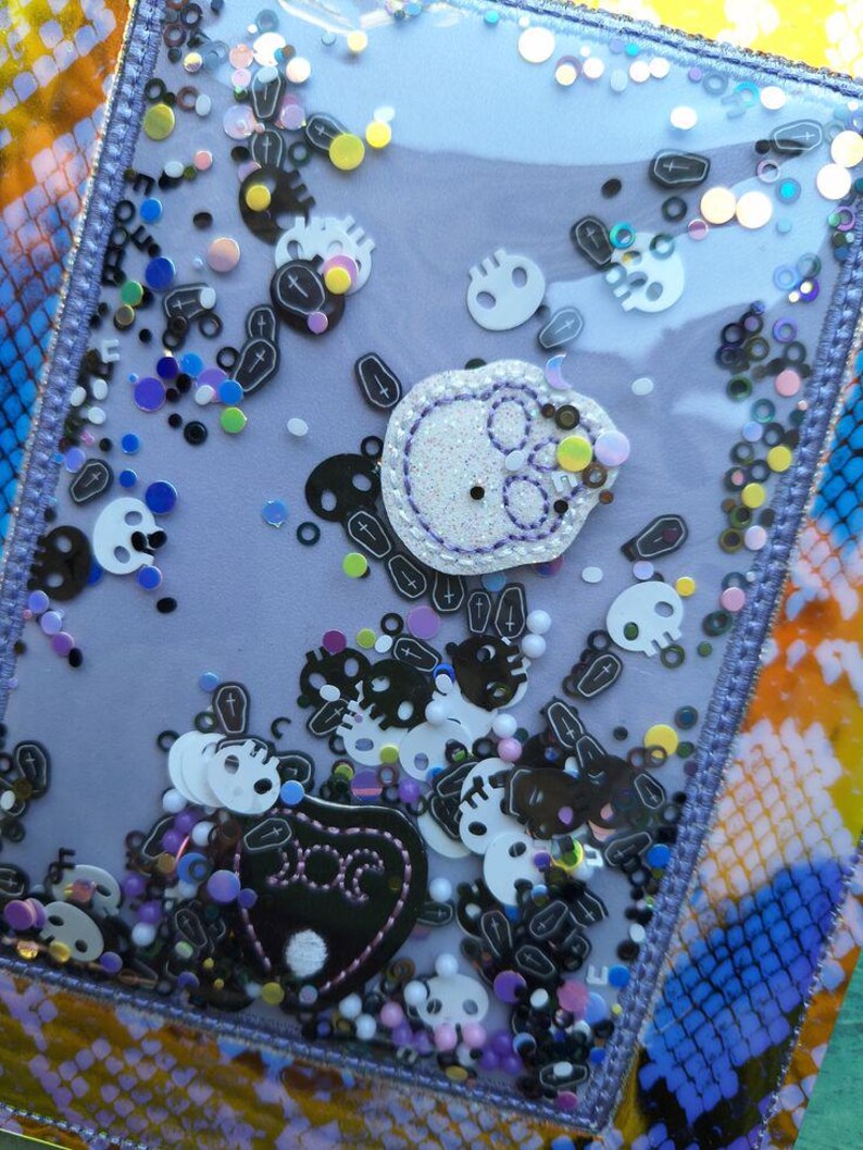 Peut inclure: Un shaker rectangulaire transparent avec un fond violet rempli de confettis en forme de cr&acirc;nes et de cercueils. Un &eacute;cusson de cr&acirc;ne blanc avec des coutures violettes est au centre. Le shaker est bord&eacute; d'une bordure violette et repose sur une surface color&eacute;e &agrave; motifs.