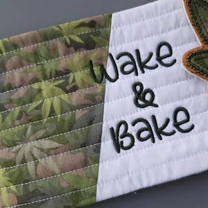 May include: A white fabric pot holder with a green camouflage pattern on one side and the words "Wake & Bake" embroidered on the other side. A green leaf-shaped patch is sewn on the white side.