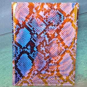 Peut inclure: Un portefeuille rectangulaire avec un motif peau de serpent dans les tons orange, bleu et blanc. Le portefeuille a une finition brillante et est cousu sur les bords. Le motif imite les &eacute;cailles d'un serpent.