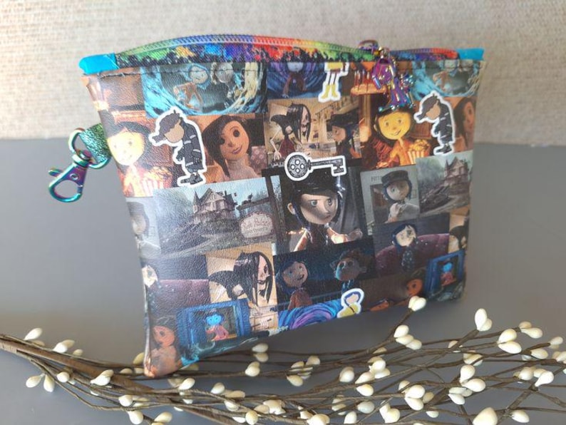 May include: A black and white patterned zippered pouch with a variety of images from the movie "Coraline". The pouch has a silver zipper and a silver key charm.