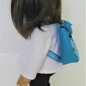 May include: A doll wearing a white shirt, black pants, and a blue and white striped backpack with a blue bow.