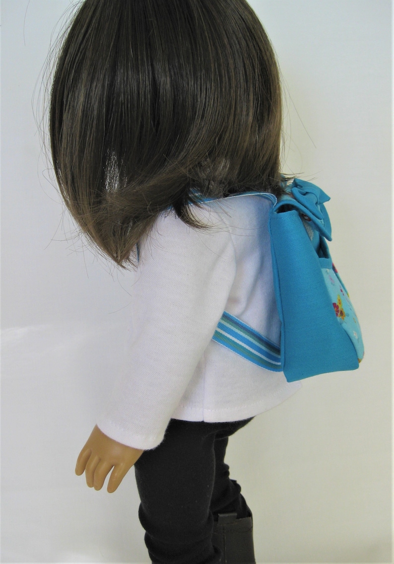 May include: A doll wearing a white shirt, black pants, and a blue and white striped backpack with a blue bow.