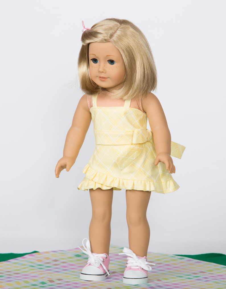 K&ouml;nnte beinhalten: Puppe mit blondem Haar und blauen Augen, die ein gelbes kariertes Kleid mit R&uuml;schenrock tr&auml;gt. Die Puppe tr&auml;gt au&szlig;erdem rosa und wei&szlig;e Turnschuhe. Eine rosa Schleife befindet sich im Haar der Puppe.