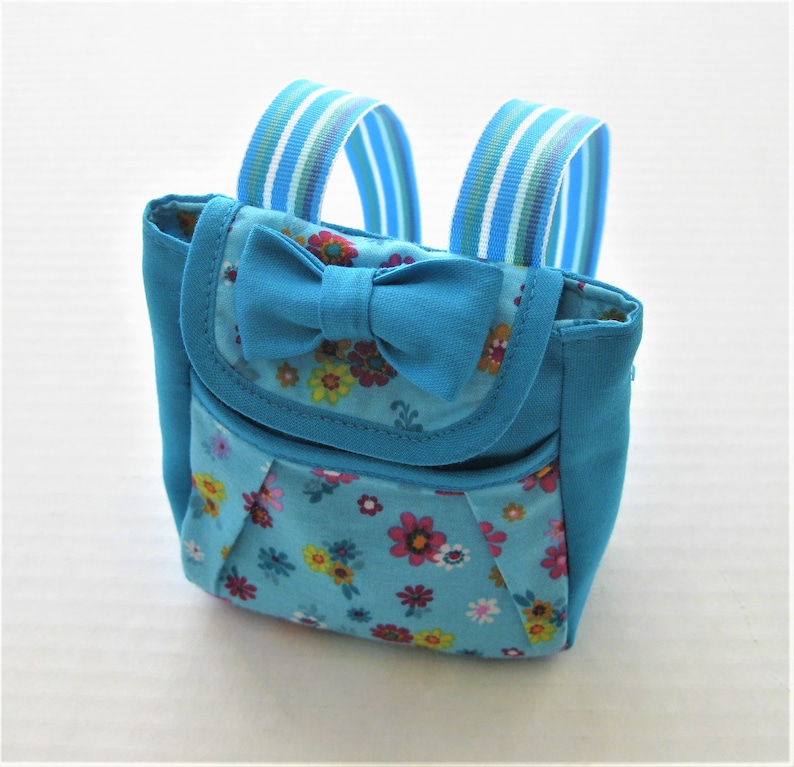May include: A small blue backpack with a floral print and a blue bow. The backpack has two adjustable straps.