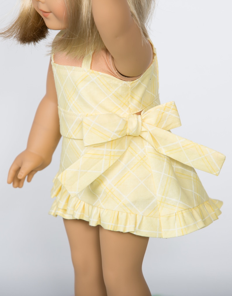 K&ouml;nnte beinhalten: Ein gelbes Kleid mit Karomuster und Schleife am R&uuml;cken. Das Kleid hat einen quadratischen Ausschnitt und einen R&uuml;schensaum. Der Stoff hat ein wei&szlig;es Gittermuster auf gelbem Hintergrund. Das Kleid ist f&uuml;r eine Puppe bestimmt.