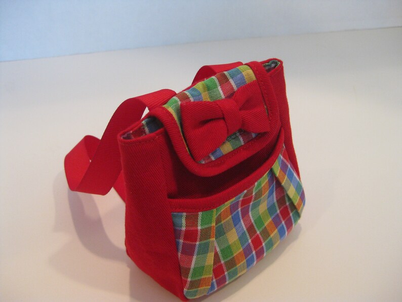 May include: A small red and multi-colored plaid backpack with a red bow on the front.