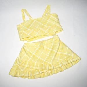 K&ouml;nnte beinhalten: Ein zweiteiliges gelbes Outfit mit Karomuster. Das Oberteil ist ein Crop-Top mit Tr&auml;gern. Der Rock ist ein Minirock mit R&uuml;schensaum. Das Outfit ist aus leichtem Stoff gefertigt und f&uuml;r warmes Wetter geeignet.