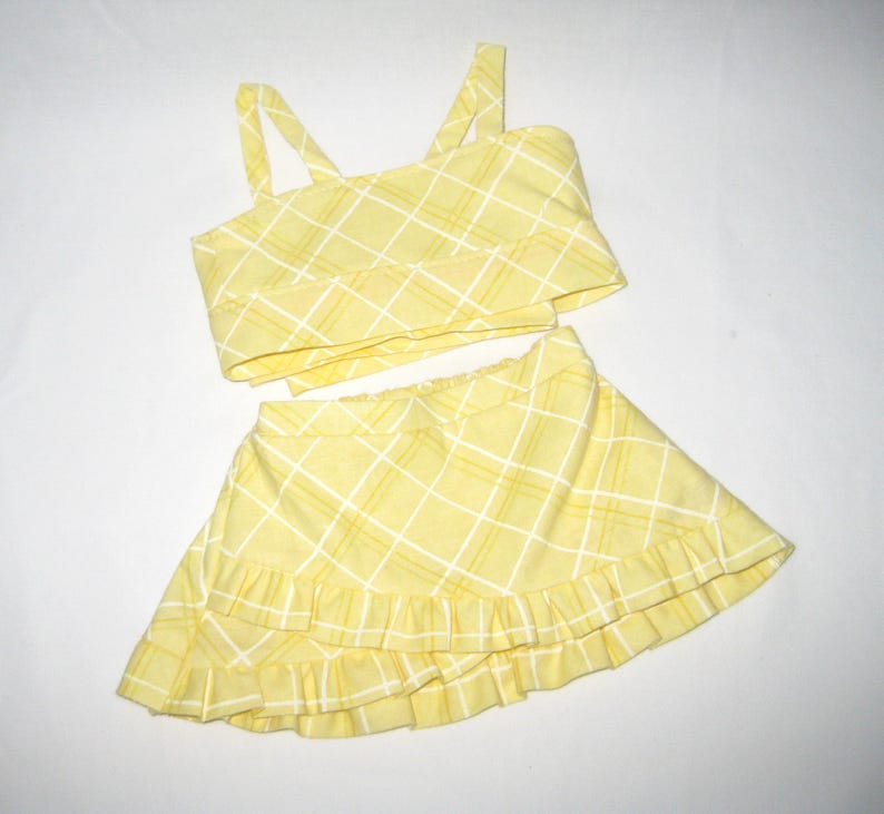 K&ouml;nnte beinhalten: Ein zweiteiliges gelbes Outfit mit Karomuster. Das Oberteil ist ein Crop-Top mit Tr&auml;gern. Der Rock ist ein Minirock mit R&uuml;schensaum. Das Outfit ist aus leichtem Stoff gefertigt und f&uuml;r warmes Wetter geeignet.