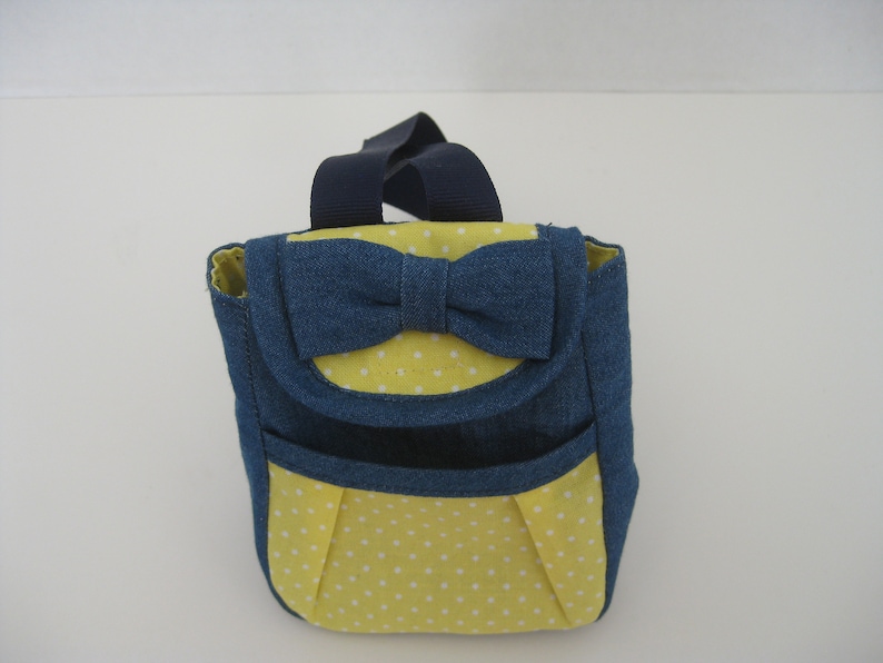 May include: A small blue denim backpack with a yellow polka dot lining and a blue denim bow on the front.