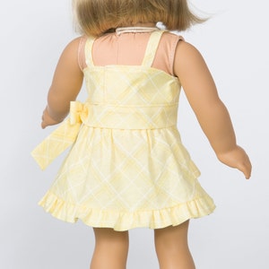 K&ouml;nnte beinhalten: Gelb kariertes Puppenkleid mit Schleife in der Taille und R&uuml;schensaum. Das Kleid hat einen quadratischen Ausschnitt und Spaghettitr&auml;ger. Das blonde Haar der Puppe ist sichtbar.
