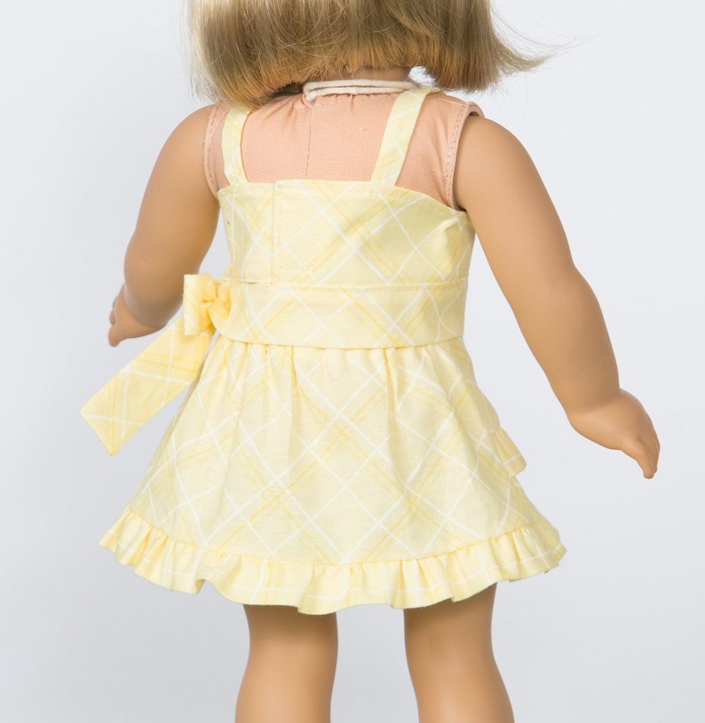 K&ouml;nnte beinhalten: Gelb kariertes Puppenkleid mit Schleife in der Taille und R&uuml;schensaum. Das Kleid hat einen quadratischen Ausschnitt und Spaghettitr&auml;ger. Das blonde Haar der Puppe ist sichtbar.