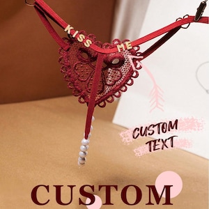 Könnte beinhalten: Burgunderfarbener String mit herzförmigem Spitzendesign. Der String hat den Text "KISS ME" in goldfarbenen Buchstaben und eine Perlenkette. Das Bild enthält auch die Wörter "CUSTOM TEXT" und "CUSTOM".