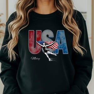 Puede incluir: Sudadera negra con la palabra "USA" en letras de bloque rojas y azules. En el centro hay un gráfico de una patinadora artística sosteniendo una bandera estadounidense. El nombre "Mary" está impreso debajo de la palabra "USA".