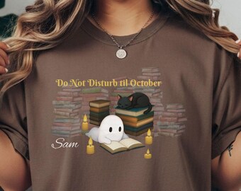 Op de afbeelding: Een bruin t-shirt met een afbeelding van een witte geest die een boek leest, een zwarte kat die op een stapel boeken slaapt en verschillende brandende kaarsen. De tekst luidt "Do Not Disturb til October" en de naam "Sam" staat in witte letters.