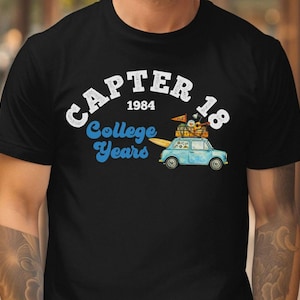 Puede incluir: Camiseta negra con un diseño gráfico que presenta el texto "CAPTER 18 1984 College Years". Un coche azul claro con equipaje y una tabla de surf también está en el diseño. La camiseta es de cuello redondo.