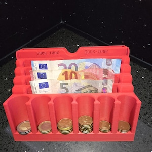 Może przedstawiać: Czerwony plastikowy organizer na pieniądze z trzema pionowymi przegródkami na banknoty i sześcioma cylindrycznymi otworami na monety. Przegródki na banknoty są oznaczone napisem 500-100 euro. Wewnątrz znajdują się banknoty i monety euro.
