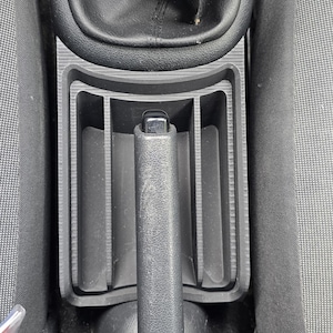 Op de afbeelding: Zwarte auto console met versnellingspook, handrem en opbergvakken. De handrem staat omhoog. De console is gemaakt van zwart plastic en is omgeven door grijze stof.