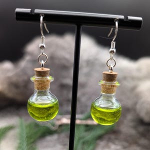 May include: A pair of glass potion bottle earrings with a green liquid inside. The earrings have cork stoppers and silver-colored hooks. The earrings are displayed on a black stand against a blurred background.