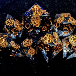 May include: Set of seven polyhedral dice with a steampunk theme. The dice are clear with a dark blue interior, gold numbers, and embedded gold-colored gears. The dice are arranged on a black surface.