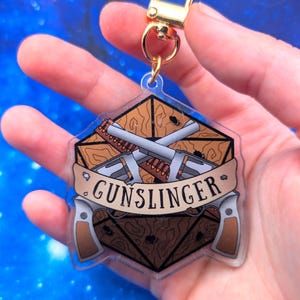 May include: A clear acrylic keychain shaped like a twenty-sided die featuring a brown wood-grain pattern, two crossed silver revolvers, and a tan banner across the center with the black text GUNSLINGER, attached to a gold-toned metal clasp.