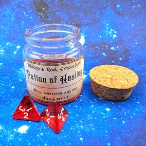 Op de afbeelding: Een doorzichtige glazen pot gevuld met rode vloeistof, met een etiket waarop staat Bishop & Rook Emporium Potion of Healing, Heal whatever ails you! Heals 2d4 + 2. Naast de pot liggen een ronde kurk en twee doorzichtige rode vierzijdige dobbelstenen.