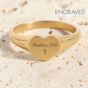 Peut inclure: Une bague dorée avec une face en forme de cœur gravée du texte Matthew 19:6 et d'un petit symbole de croix. L'anneau présente trois lignes horizontales gravées de chaque côté, et le mot ENGRAVED est visible dans le coin supérieur droit.