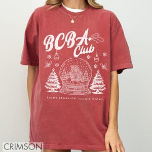 Puede incluir: Una camiseta de color rojo con un diseño gráfico blanco. El diseño incluye el texto "BCBA Club" encima de una ilustración de globo de nieve con una casa y árboles de Navidad. La frase "EVERY BEHAVIOR TELLS A STORY" está debajo.
